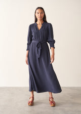 A woman modeling a navy blue midi dress with long sleeves, a smocked chest detail, and a matching fabric tie belt, paired with brown platform sandals against a neutral studio backdrop.