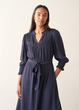 A woman modeling a navy blue midi dress with a smocked yoke, long sleeves with ruffled cuffs, and a matching fabric tie belt, set against a plain light beige background.