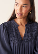 A close-up shot of a woman wearing a dark navy blue blouse with a smocked, textured yoke and a split neckline.