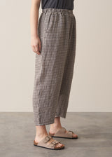 Woman wearing grey checked cropped linen barrel leg trousers with a grey vest top and neutral sandals, against a beige background