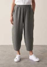 A woman wears a white linen shirt and grey linen cropped barrel-leg trousers with a pair of white trainers, against a beige background