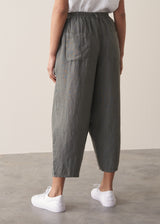 Woman wears a white linen shirt and grey linen cropped barrel leg trousers with white trainers, against a beige background