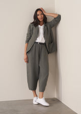 A woman stands against a beige background, dressed in a grey linen jacket over a white linen shirt, with matching linen cropped barrel leg trousers and white trainers