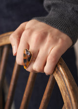 Close up of woman's hand resting on a wooden char, wearing a gold plated, oversized, brown tortoiseshell heart ring