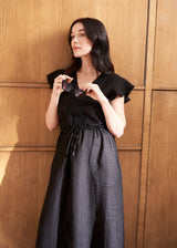 Woman wearing a black linen midi dress with drawstring waist and embroidery detail against a wooden background
