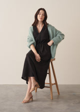 A women model wearing a black linen midi dress with a drawstring waist and embroidery detail, layered over a mint green chunky knit cardigan, against a beige background