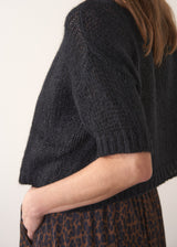 Side view of woman wearing a black sparkle knit short sleeve sweater and a leopard print skirt on a plain background