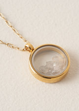 Close-up of a long, delicate gold chain necklace with a circular pendant featuring grey crystals on a neutral beige background