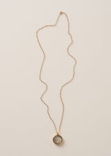 A long, delicate gold chain necklace with a circular pendant featuring grey crystals on a neutral beige background