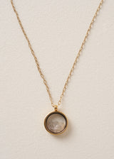 Close-up of a long, delicate gold chain necklace with a circular pendant featuring grey crystals on a neutral beige background