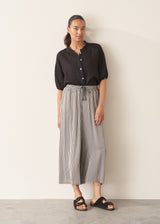 Woman wearing a black linen top and striped, cropped satin trousers against plain background