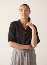 Woman wearing a black linen top and striped, cropped satin trousers against plain background