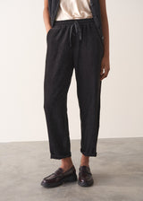 Woman wearing a black waistcoat and black slimfit drawstring trousers against a white wall.