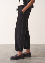 Woman wearing a black waistcoat and black slimfit drawstring trousers against a white wall.