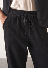 Woman wearing a black waistcoat and black slimfit drawstring trousers against a white wall.