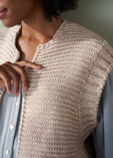 Close up of a woman wearing a beige chunky knit vest over a pale blue shirt and dark green trousers with a neutral background