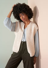 Woman wearing a beige knitted vest over a pale blue shirt and dark green trousers leaning against a neutral wall