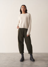 Woman wearing a round neck oatmeal knit sweater with green trousers and black boots, against a plain background