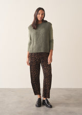 Woman wearing a sage green sleeveless sweater over a long sleeve top and dark brown leopard print trousers against a plain background.