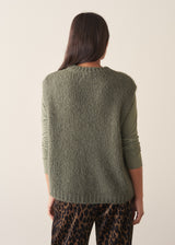 Woman wearing a sage green sleeveless sweater over a long sleeve top and dark brown leopard print trousers against a plain background.
