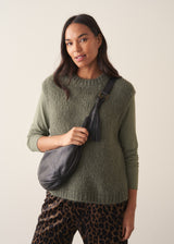 Woman wearing a sage green sleeveless sweater over a long sleeve top and dark brown leopard print trousers against a plain background.