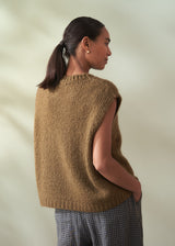 Woman wearing a khaki green knitted vest with check trousers facing a neutral background