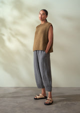 Person wearing a khaki green sleeveless sweater and checkered trousers standing against a neutral background