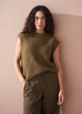 Woman in a sleeveless knitted tank and khaki trousers, leaning against a neutral background.