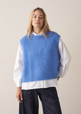 Woman wearing blue knitted sleeveless sweater, a white shirt and dark blue jeans