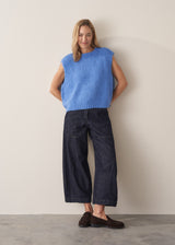 Woman models a blue knitted sleeveless tank and dark blue jeans with loafers, set against a beige background
