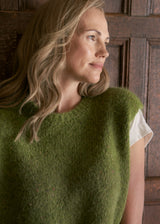 A woman with blonde hair wearing a fuzzy olive green sleeveless sweater vest over a white short-sleeve top, standing in front of a dark wooden paneled wall.