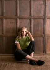 A woman sits on a wooden floor in front of a dark wood paneled wall, wearing a textured olive green knit sweater vest over a white t-shirt, black cropped pants, and black sneakers.