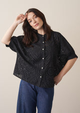 Woman wearing black broderie anglaise boxy shirt with blue jeans against a beige background