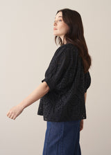Woman wearing black broderie anglaise boxy shirt with blue jeans against a beige background