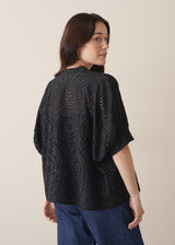 Woman wearing black broderie anglaise boxy shirt with blue jeans against a beige background