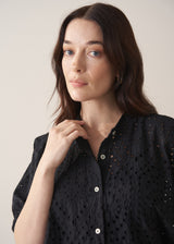 Close up of woman wearing black broderie anglaise boxy shirt against a beige background
