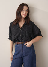 Woman wearing black broderie anglaise boxy shirt with belted blue jeans against a beige background