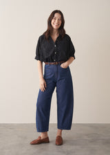 Woman wearing black broderie anglaise boxy shirt with blue jeans, styled with brown loafers against a beige background