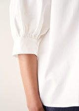 A woman wearing a white-button down cotton shirt with three-quarter puff sleeves and a relaxed fit, with blue denim jeans.