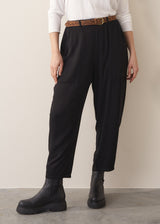 A woman wearing a white top and black barrel leg trousers with leopard print belt and chunky boots against a plain background