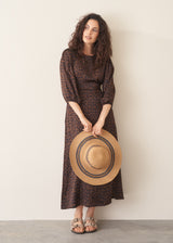 Woman in a brown patterned dress holding a large straw hat against a white wall.