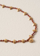 Close up detail of necklace with plum crystal beads, gold discs, and a hammered heart pendant, on a beige background