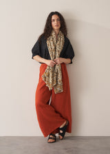 Women leant against a wall wearing a pair of full length wide leg linen trousers in burnt orange, paired with a black broderie top and sandals