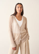 A woman wearing a soft knit cardigan in a light beige colour, worn over a white vest top and paired with matching beige trousers.