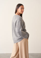 A woman wearing a grey knit cardigan over a white top and beige trousers, photographed against a plain studio background.