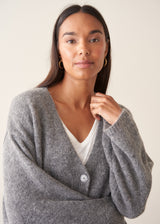 A woman wearing a grey knit cardigan over a white top and beige trousers, photographed against a plain studio background.