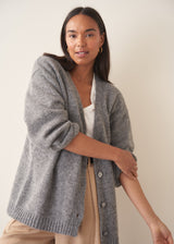 A woman wearing a grey knit cardigan over a white top and beige trousers, photographed against a plain studio background.