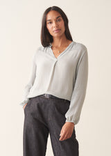 A woman wearing a light blue v-neck blouse with dark jeans against a plain background