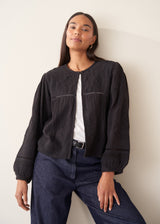 A woman wearing a black embroidered textured cropped jacket over a white top and dark blue jeans.