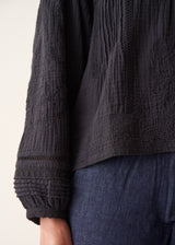 A woman wearing a black embroidered textured cropped jacket over a white top and dark blue jeans.